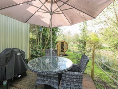 An outdoor patio area with a glass table, four wicker chairs, an umbrella, and a covered grill at Damsel Shepherds Hut in Badwell Ash