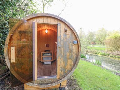 A wooden barrel sauna with a glass door next to a pond and greenery at Damsel Shepherds Hut in Badwell Ash