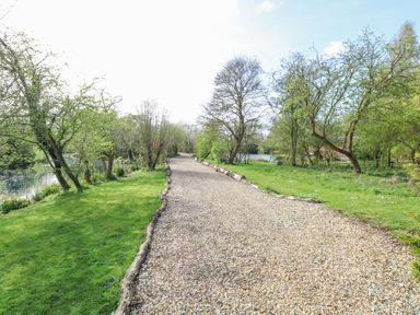 A gravel path with trees and grass on either side near a body of water at Damsel Shepherds Hut in Badwell Ash