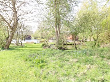 A grassy garden area with trees and a wooden swing bench near a lake with cabins in the background at Damsel Shepherds Hut in Badwell Ash