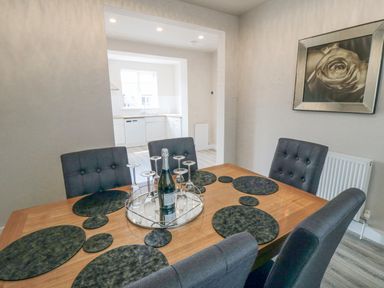 A dining room with a wooden table set with a bottle and upside down glasses and gray chairs next to an open kitchen at Ocean Spray in Scarborough