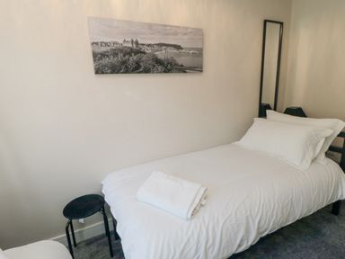 A bedroom with a single bed white bedding a folded towel a black stool a tall mirror and a black and white wall picture at Ocean Spray in Scarborough