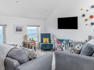 A living room with grey sofas a blue chair a wall-mounted television and windows showing the sea at Loe Cottage in Porthleven