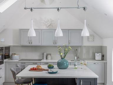 A kitchen with grey cabinets white countertops a kitchen island with chairs flowers bread and wine at Loe Cottage in Porthleven