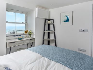 A bedroom with a bed a window desk with a lamp and flowers and a corner shelf with a painting on the wall at Loe Cottage in Porthleven