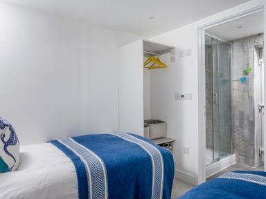 A bedroom with two single beds covered in blue striped blankets and a pillow on one bed a small open closet with yellow hangers and storage baskets and an en suite bathroom with a glass shower enclosure at Loe Cottage in Porthleven