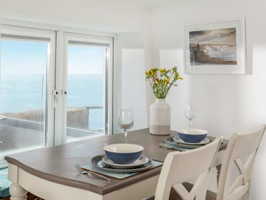 A dining table set for two with plates bowls glasses and a vase of flowers by a window with ocean view at Loe Cottage in Porthleven