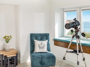 A corner with a blue chair and a pillow next to a set of nested tables with a vase of flowers and a telescope by a window with a sea view at Loe Cottage in Porthleven