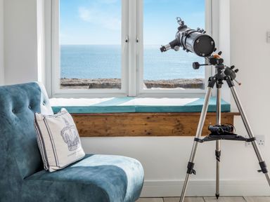 A telescope on a tripod next to a blue chair with a cushion by a window overlooking the sea at Loe Cottage in Porthleven