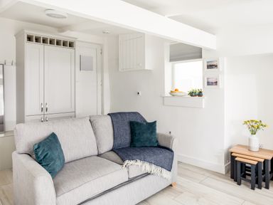 A living room with a gray sofa with blue pillows and blanket a white cabinet a small set of nesting tables with flowers and framed pictures on the wall at Loe Cottage in Porthleven