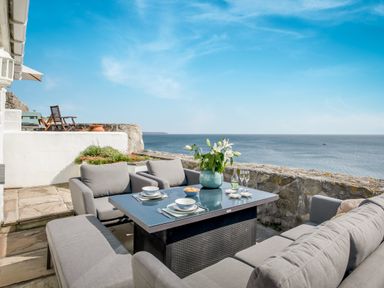 An outdoor patio with gray cushioned seating and a table set with dishes and flowers overlooking the sea at Loe Cottage in Porthleven