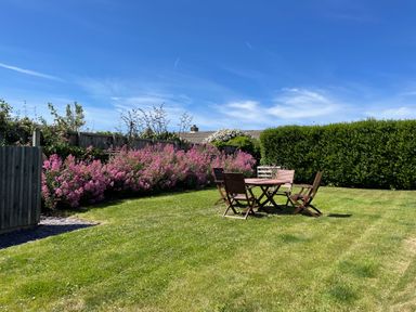 A garden with a wooden table and four chairs on grass with pink flowers and green hedge at The Old Boathouse in Trefin near St Davids