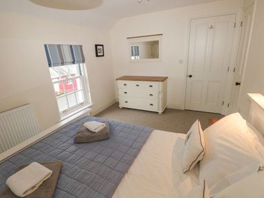 A bedroom with a bed covered with a blue quilt and white pillows a white dresser with a wooden top a wall mirror a window with striped blinds and a closed door at The Old Boathouse in Trefin near St Davids