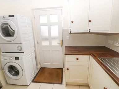 A laundry room with stacked washer and dryer next to a door and countertop with sink at The Old Boathouse in Trefin near St Davids