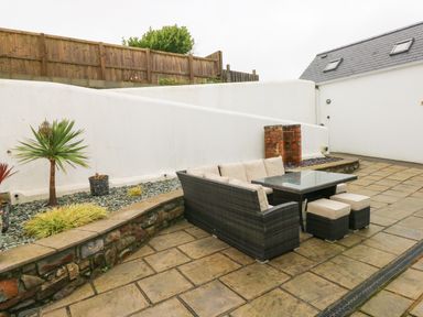An outdoor patio area with a wicker sofa, glass table, two ottomans, potted plants, stone wall, and a wooden fence at The Old Boathouse in Trefin near St Davids