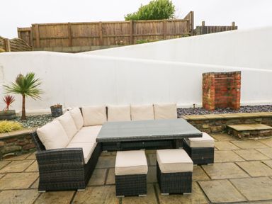 An outdoor patio area with a cushioned wicker corner sofa table and stools with a brick barbecue and wooden fence at The Old Boathouse in Trefin near St Davids