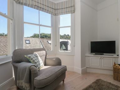 A sitting area with a gray armchair and a television on a cabinet near a corner window at 21 Passage Street formerly Panacea in Fowey