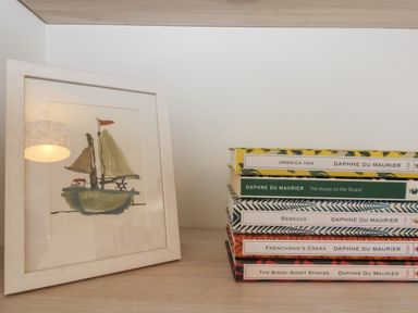 A framed painting of a sailboat next to a stack of books by Daphne du Maurier on a wooden surface at 21 Passage Street formerly Panacea in Fowey