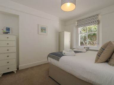 A bedroom with a bed, a white chest of drawers, a white wardrobe, and a window with a patterned blind at 21 Passage Street formerly Panacea in Fowey