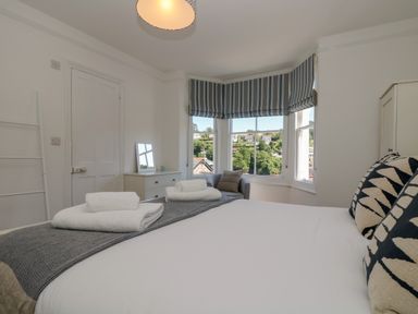 A bedroom with a bed, folded towels, patterned pillows, a chair by bay windows with striped blinds, a dresser with a mirror at 21 Passage Street formerly Panacea in Fowey