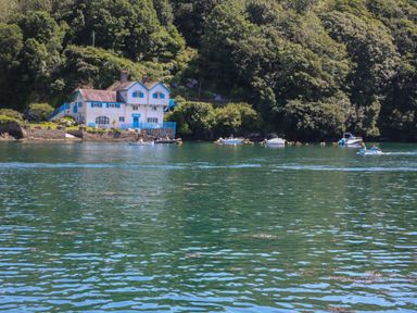A white house with blue trim by a body of water with several small boats and trees on the shore at 21 Passage Street formerly Panacea in Fowey