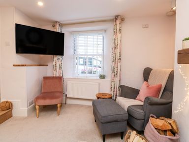 A living room with a gray armchair and ottoman a pink chair a wall-mounted television and a window with curtains at Pilchard Cottage in Dawlish