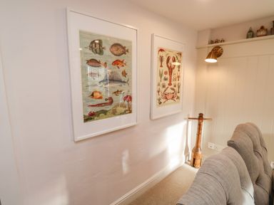 Two framed fish and sea creature illustrations on a wall above a gray sofa with wooden sculpture and wall light at Pilchard Cottage in Dawlish