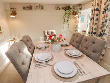 A dining table set with plates and cutlery surrounded by six upholstered chairs with a vase of red flowers at Pilchard Cottage in Dawlish