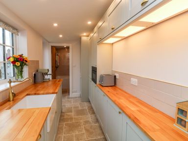 A narrow kitchen with wooden countertops a sink a kettle a toaster flowers on the windowsill and cabinets on both sides at Pilchard Cottage in Dawlish