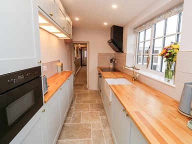 A galley kitchen with wooden countertops, cabinets, a built-in oven, a sink under a window, and a vase of flowers on the windowsill at Pilchard Cottage in Dawlish
