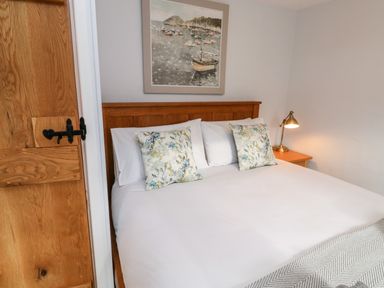 A bedroom with a wooden bed with white bedding and floral pillows a wooden door a framed painting and a bedside table with a lamp at Pilchard Cottage in Dawlish