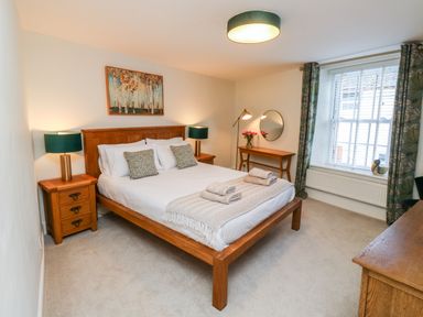 A bedroom with a wooden bed double bedside tables lamps a wooden dressing table with a mirror a window and curtains at Pilchard Cottage in Dawlish