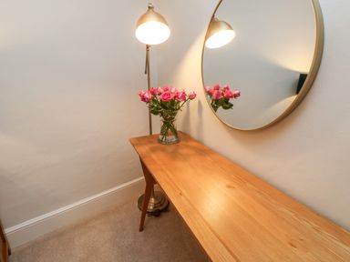 A wooden console table with a vase of pink flowers a round wall mirror and a tall floor lamp at Pilchard Cottage in Dawlish