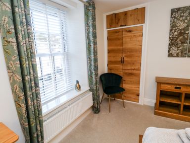 A room with a window with floral curtains a black chair wooden cupboards and a wooden shelving unit at Pilchard Cottage in Dawlish
