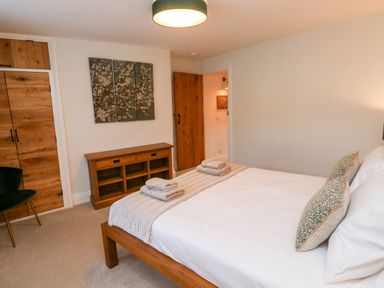 A bedroom with a wooden bed, pillows, folded towels on the bed, a wooden cabinet, a chair, and a triptych painting on the wall at Pilchard Cottage in Dawlish