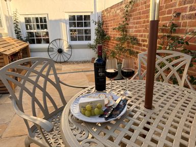 A patio table with two glasses of red wine a bottle cheese grapes and crackers with chairs on a stone floor and brick and white walls at Pilchard Cottage in Dawlish
