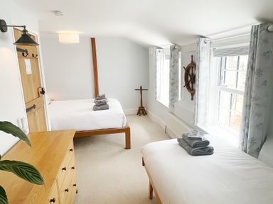 A bedroom with two beds and stacks of towels by windows with curtains and a wooden dresser at Pilchard Cottage in Dawlish