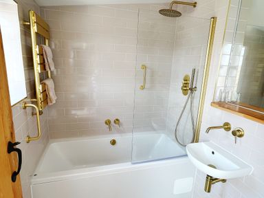 A bathroom with a bathtub shower combination towels on a rack and a sink with brass fixtures at Pilchard Cottage in Dawlish