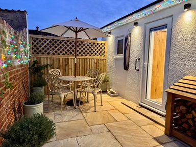 An outdoor patio with a round table and four chairs under an umbrella near a white building with string lights and potted plants at Pilchard Cottage in Dawlish