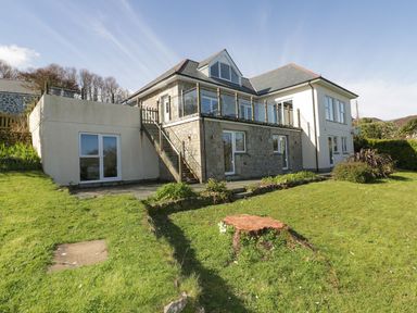 Exterior view of a two-story house with stone and white walls a green lawn and a tree stump in the garden at Lower Mellan Barn in Coverack