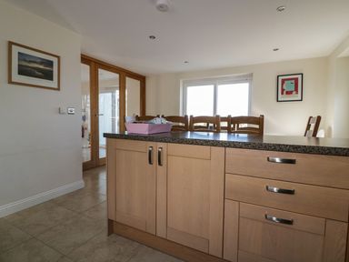 A kitchen island with wooden cabinets and a granite countertop with chairs and framed pictures on the walls at Lower Mellan Barn in Coverack