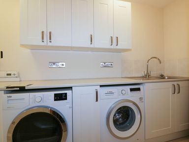 A laundry room with a washing machine and dryer under a countertop with white cabinets above and a sink at Lower Mellan Barn in Coverack