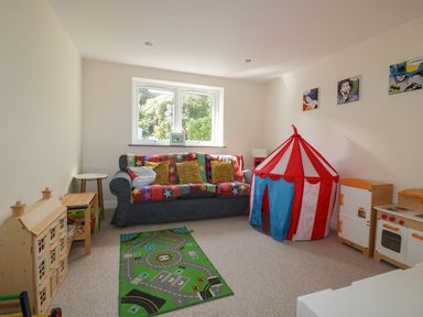 A children's playroom with a gray sofa covered by a colorful star blanket and yellow pillows a small circus tent a play kitchen a dollhouse and a green play mat on the floor at Lower Mellan Barn in Coverack