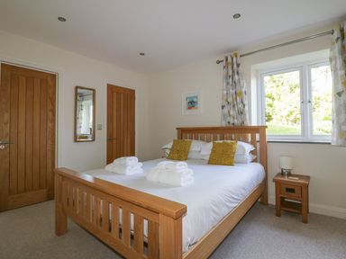 A bedroom with a wooden bed frame white bedding folded towels two wooden doors a window with curtains and a bedside table lamp at Lower Mellan Barn in Coverack