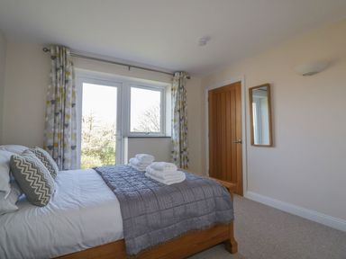 A bedroom with a wooden bed covered with a gray blanket and pillows next to a window with curtains at Lower Mellan Barn in Coverack