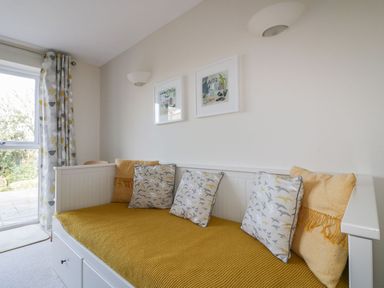 A white daybed with yellow cushions and bird-patterned pillows in a room with framed pictures and patterned curtains at Lower Mellan Barn in Coverack