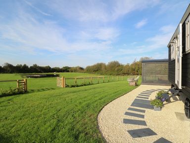 A grassy field with a fenced gate beside a path leading to a black mobile hut with stairs and potted plants at Dragonfly Shepherd's Hut Oakford near Aberaeron