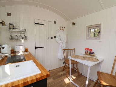 A small kitchen with wooden worktops a white sink a table with two wooden chairs and a wall with hanging robes at Dragonfly Shepherd's Hut in Oakford near Aberaeron