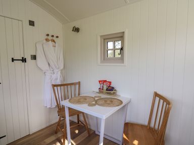 A small dining area with a white table and two wooden chairs white robes hanging on wall hooks and a small window at Dragonfly Shepherd's Hut in Oakford near Aberaeron