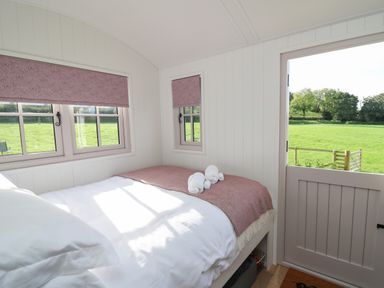 A bedroom with a bed with white sheets and pink blanket next to windows and a half open door showing a green field at Dragonfly Shepherd's Hut in Oakford near Aberaeron